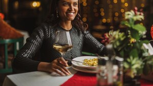 Une personne assise à une table, devant un verre de vin, une assiette de nourriture et un vase rempli de fleurs.
