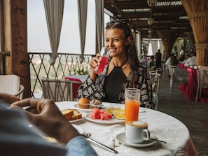 A person seated at a table with a drink, food, and a cup, in a covered outdoor dining area with other tables and chairs visible. - Pyramid Hotels & Resorts  (Bishoftu)
