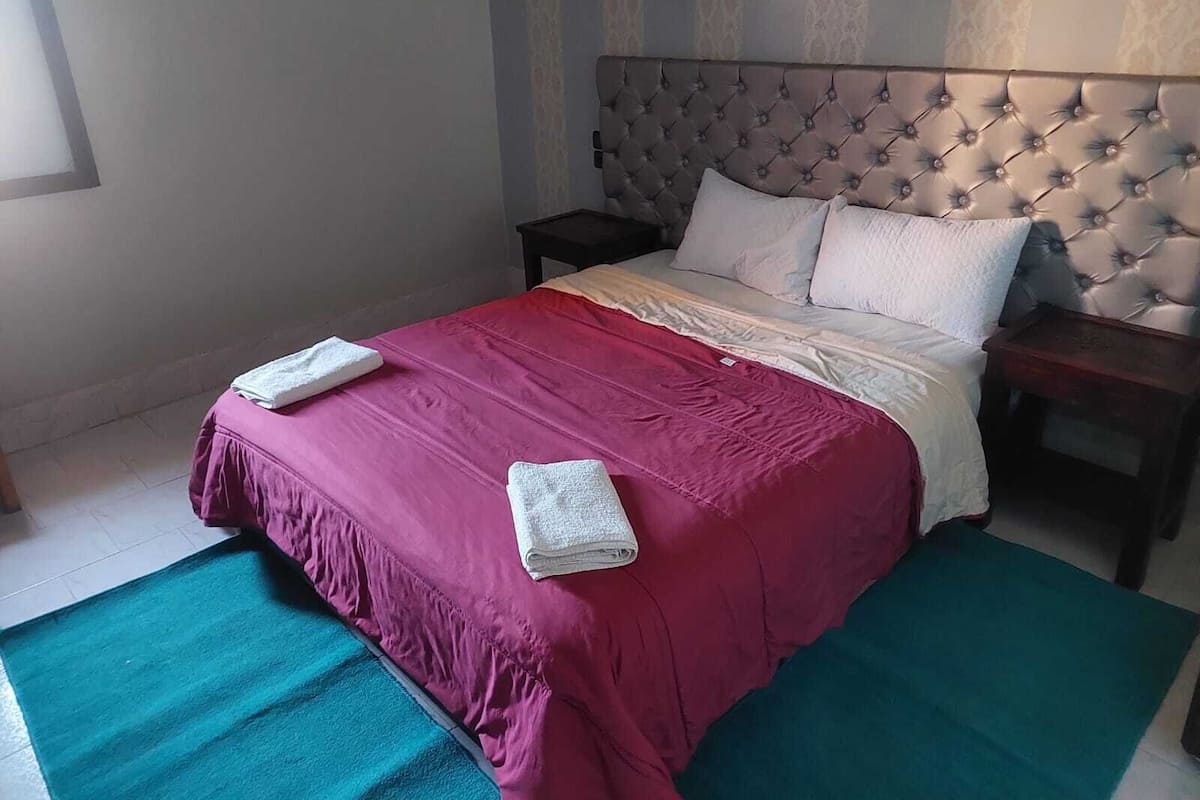 A neatly made bed with a red bedspread and white pillows, a wooden nightstand, and a teal rug.
