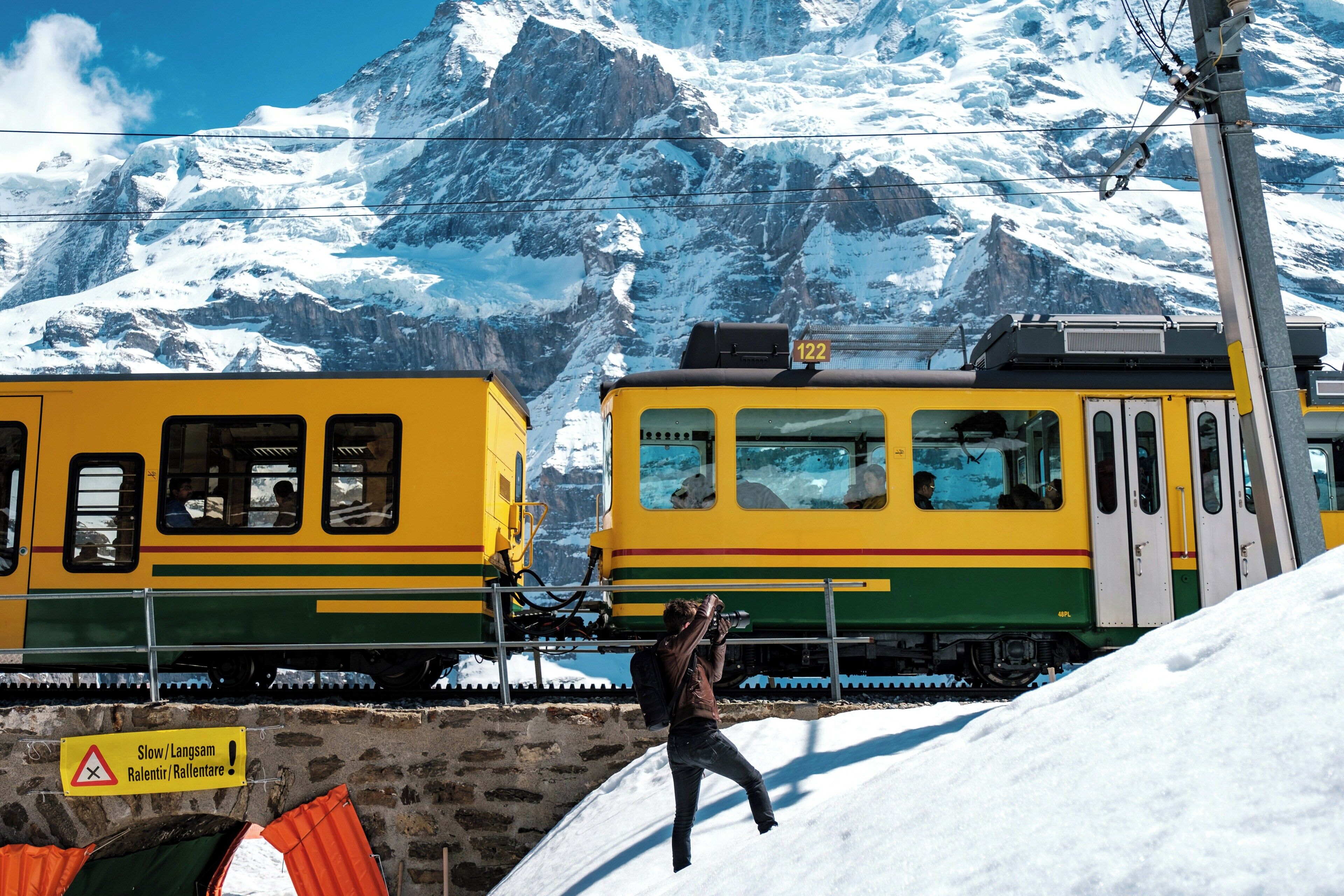 A train with a yellow and green color scheme, a person taking a photo, and a mountainous snowy background.