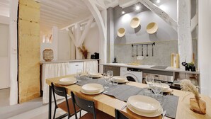 A dining area with a wooden table set for four, surrounded by chairs. A kitchen with modern appliances is visible in the background. - Elegant Apartment -2br-4p- Le Marais (Paris)