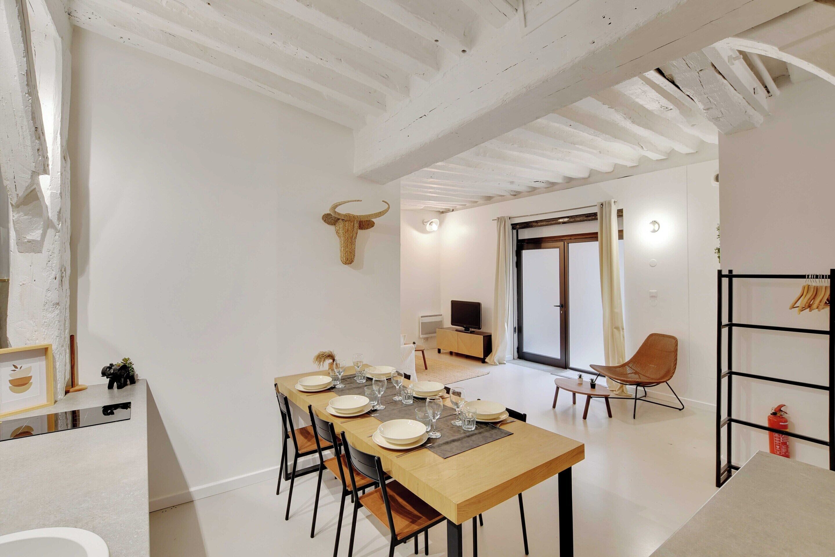 A dining area with a long wooden table set for six, surrounded by chairs. A bull's head decoration hangs on the wall, and a television is visible in the background.