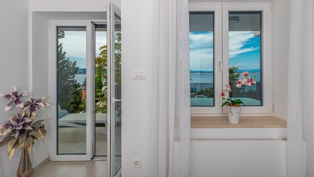 A room with a sliding glass door leading to an outdoor area, a window with a view of the landscape, and a potted plant on the windowsill. - Apartman Maxim (Crikvenica)