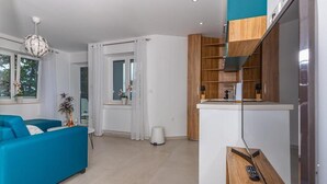 A modern living room with a blue sofa, a small table with a vase, and a kitchen area with wooden cabinets and a countertop. - Apartman Maxim (Crikvenica)