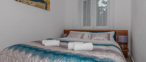 A bedroom with a bed, a window with curtains, a nightstand with a lamp, and a painting on the wall.