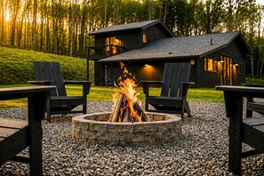 A cabin with a stone fire pit surrounded by Adirondack chairs, set against a forest backdrop.
