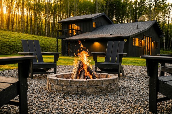 A cabin with a stone fire pit surrounded by Adirondack chairs, set against a forest backdrop.