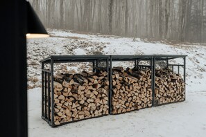 A metal structure with stacked firewood in a snowy outdoor setting. - Onyx Ridge · Breathtaking Views · 14 Acres (Jeffersonville)
