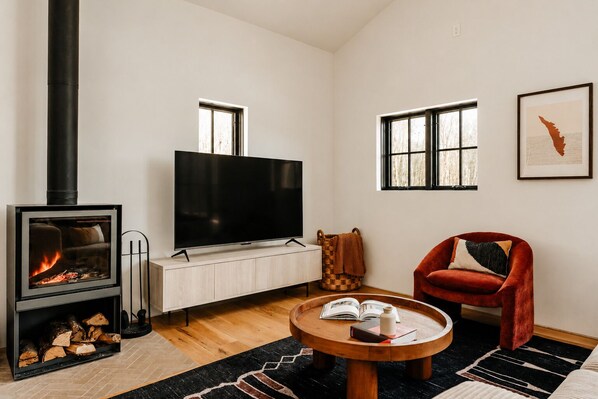 A living room with a fireplace, a flat-screen TV, a red chair, and a round wooden table with books and a bottle.