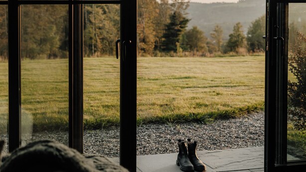 A view through large glass doors of a grassy field and trees, with a pair of boots on a rug in the foreground. - Hot Tub, Fire Pit, Views & 14 Acres | Onyx Ridge (Jeffersonville)