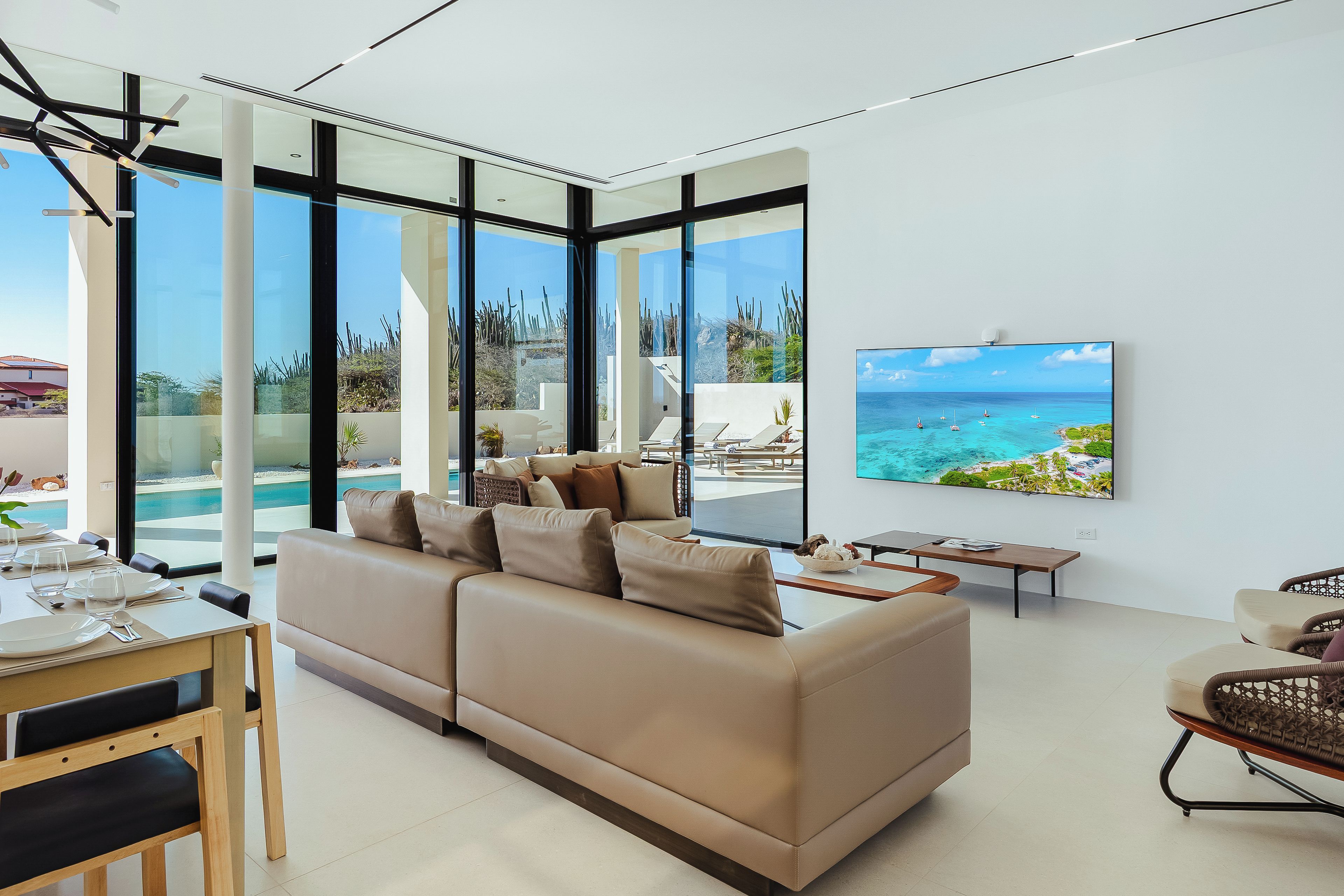 A modern living room with a large sofa, a dining area, and a TV displaying a beach scene.