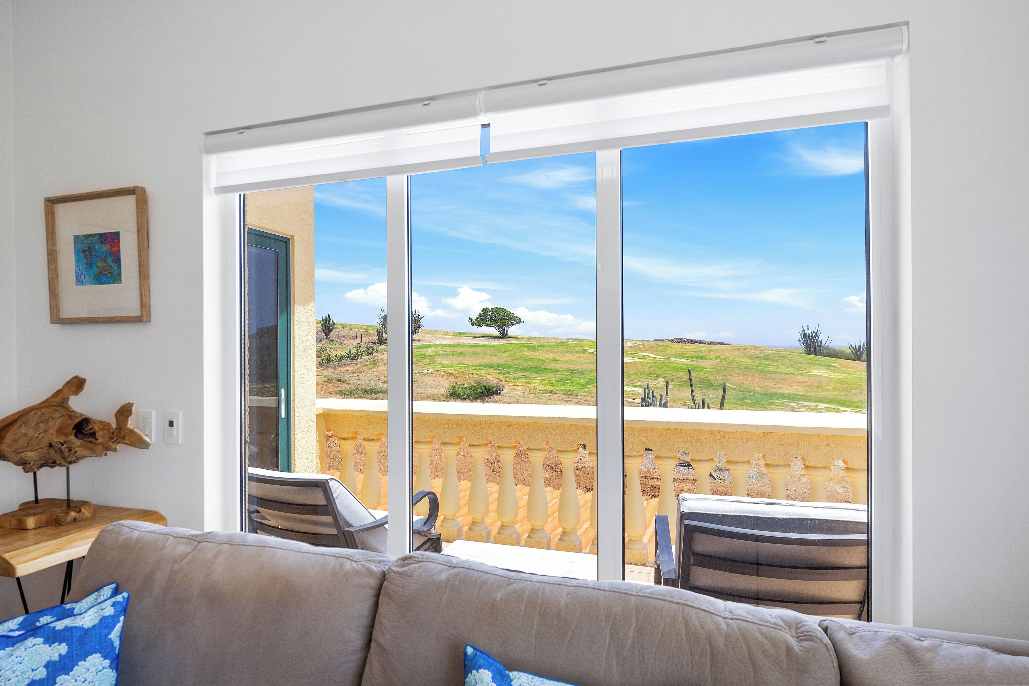 A living room with a large sliding glass door leading to a balcony with a view of a grassy landscape and a tree.