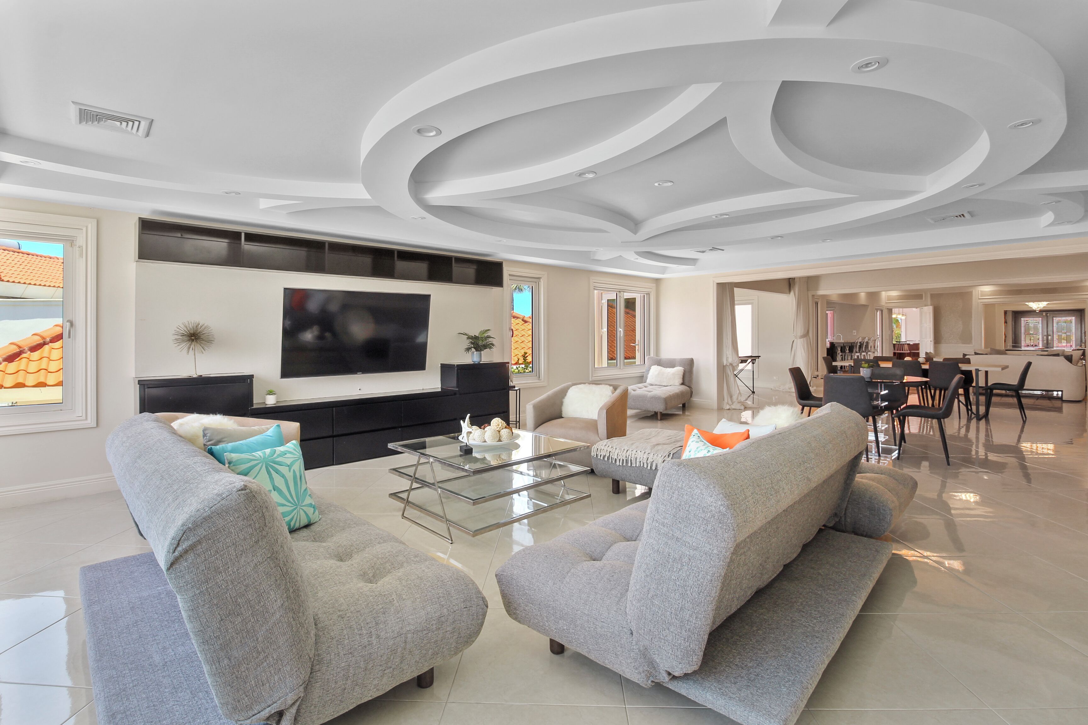 A modern living room with a flat-screen TV, grey sofas, a glass coffee table, and a dining area in the background.