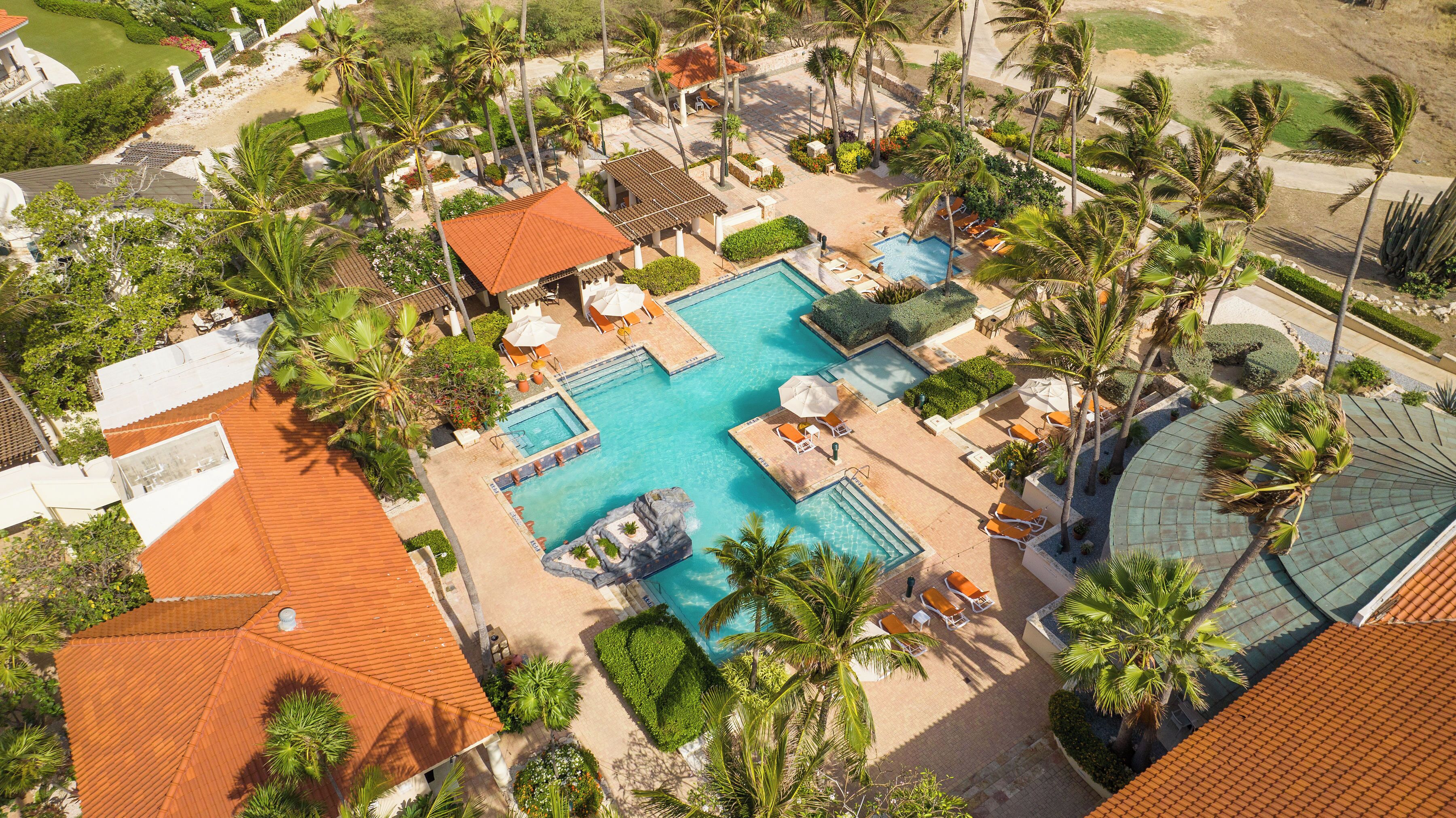 Luchtbeeld van een resort met een centraal gelegen zwembad, meerdere loungegedeeltes en palmbomen.