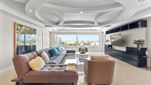Un salón espacioso con un sofá modular grande, una mesa de centro redonda y un sillón cómodo. Un televisor de pantalla plana está montado en la pared, y hay un ventanal que ofrece vistas al exterior.