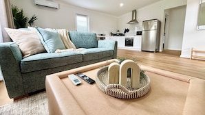 A modern living room with a grey sofa, a beige armchair, and a woven coffee table. A kitchen with white cabinets and stainless steel appliances is visible in the background. - Joondalup Hidden Villa Gemma w Pool! (Heathridge)