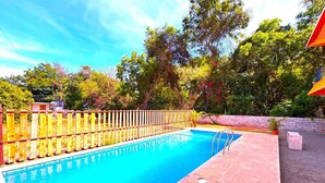 A rectangular swimming pool with a wooden fence, surrounded by a paved area and trees. - De Capital Hotel (Vanur)
