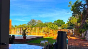 A view from a room with a wooden table, a potted plant, and a metal container. Outside, there is a wooden fence, a small lawn, and trees. - De Capital Hotel (Vanur)