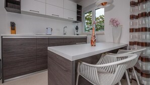 A modern kitchen with dark wood cabinets, a white countertop, and a bar area with white chairs. - Apartman Konstantin (Crikvenica)