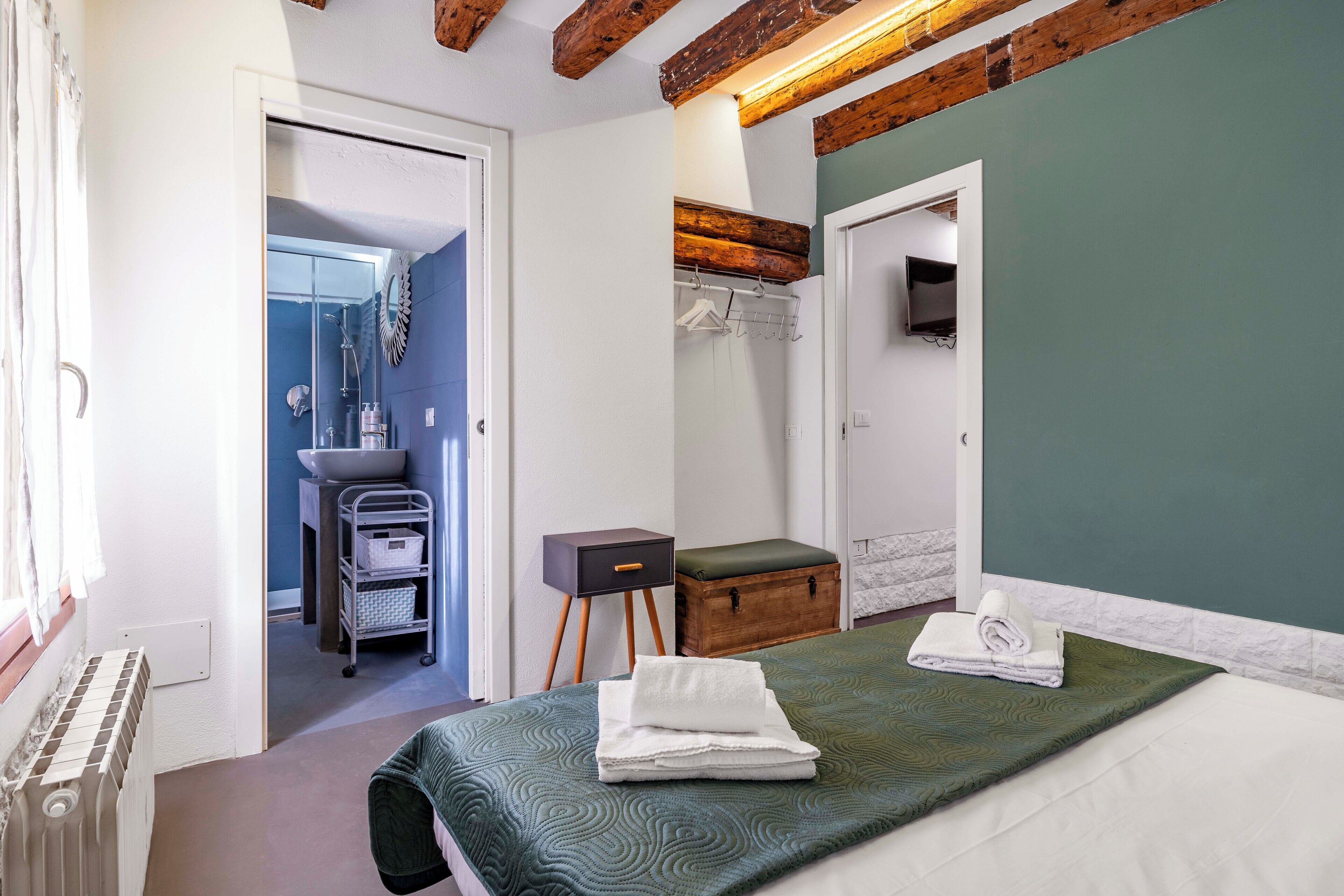 Une chambre à coucher avec un lit, une commode en bois, une petite table, un miroir et une salle de bain visible à travers une porte ouverte.