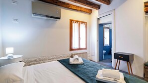 Un dormitorio con cama, una mesita de noche con lámpara, un radiador, una ventana con cortinas, una pequeña mesita de noche y una puerta que da a otra habitación.