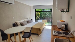 Une chambre d’hôtel moderne équipée d’un lit, d’une petite table avec des chaises, d’un coin cuisine et offrant une vue sur la verdure par la fenêtre.