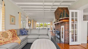 A spacious living room with a grey sectional sofa, a round coffee table, and a brick fireplace. - Waterfront, Walk to Beach! (Barnstable)