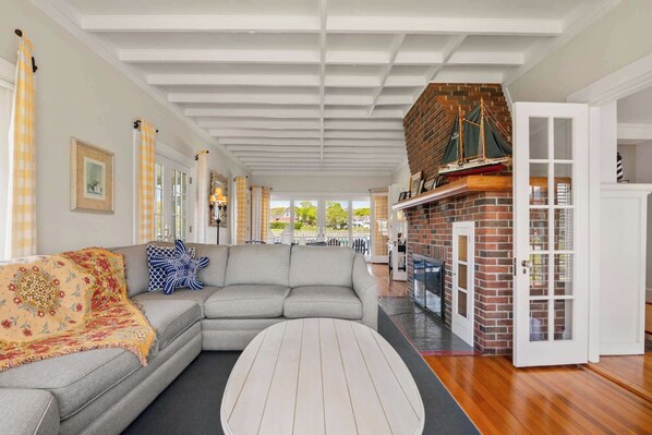 A spacious living room with a grey sectional sofa, a round coffee table, and a brick fireplace. - Waterfront, Walk to Beach! (Barnstable)
