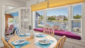 A dining area with a view of a lake and houses, featuring a wooden table set for four, a bench with cushions, and a window with a yellow awning. - Waterfront, Walk to Beach! (Barnstable)