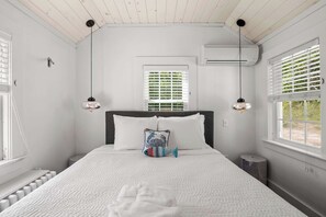 A bedroom with a bed, two windows with blinds, an air conditioning unit, and a radiator.