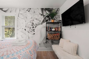 Een slaapkamer met een groot bloemenmural, een boekenkast, een televisie en een raam met jaloezieën.