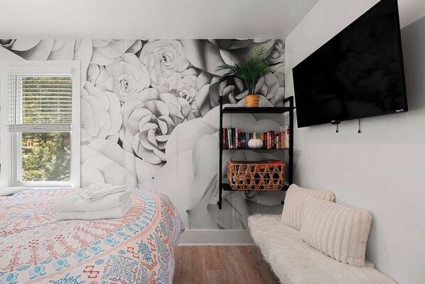 A bedroom with a large floral mural, a bookshelf, a TV, and a window with blinds.