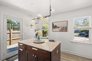 A kitchen with a marble countertop, wooden cabinets, hanging light fixtures, and a view of the outdoors through the windows. - Adorable Updated Cottage, Walk to Water! (Yarmouth)