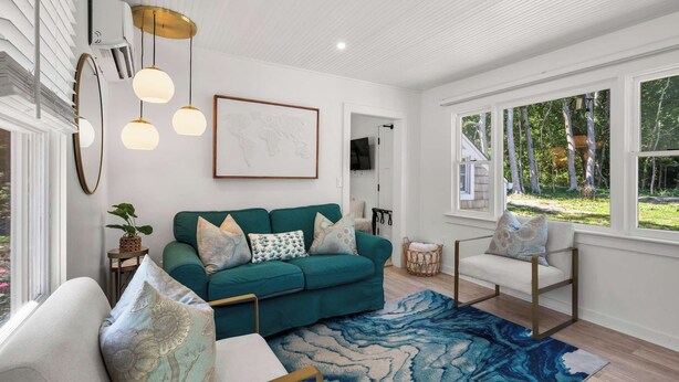 A modern living room with a teal sofa, patterned cushions, a blue and white rug, and a large window with a view of trees. - Adorable Updated Cottage, Walk to Water! (Yarmouth)