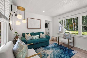 Een moderne woonkamer met een blauwgroene bank, gestreepte kussens, een blauw-wit kleed en een groot raam met uitzicht op bomen.