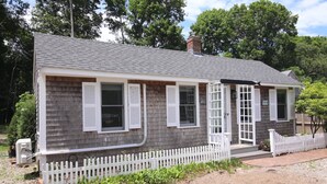 A small, single-story house with a grey shingle roof, white shutters, and a white picket fence. - Adorable Updated Cottage, Walk to Water! (Yarmouth)