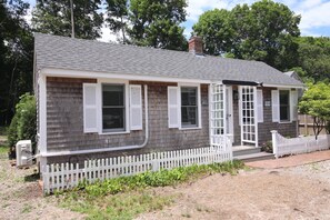 A small, single-story house with a grey shingle roof, white shutters, and a white picket fence. - Adorable Updated Cottage, Walk to Water! (Yarmouth)