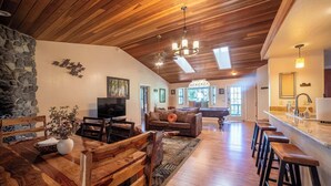 A spacious living area with a wooden ceiling, a stone wall, a dining table with chairs, a sofa, and a kitchen area with bar stools. - Sierra Grand Inside Yosemite (Wawona)