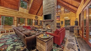A log cabin interior with a stone fireplace, leather sofas, and a large window overlooking the forest.