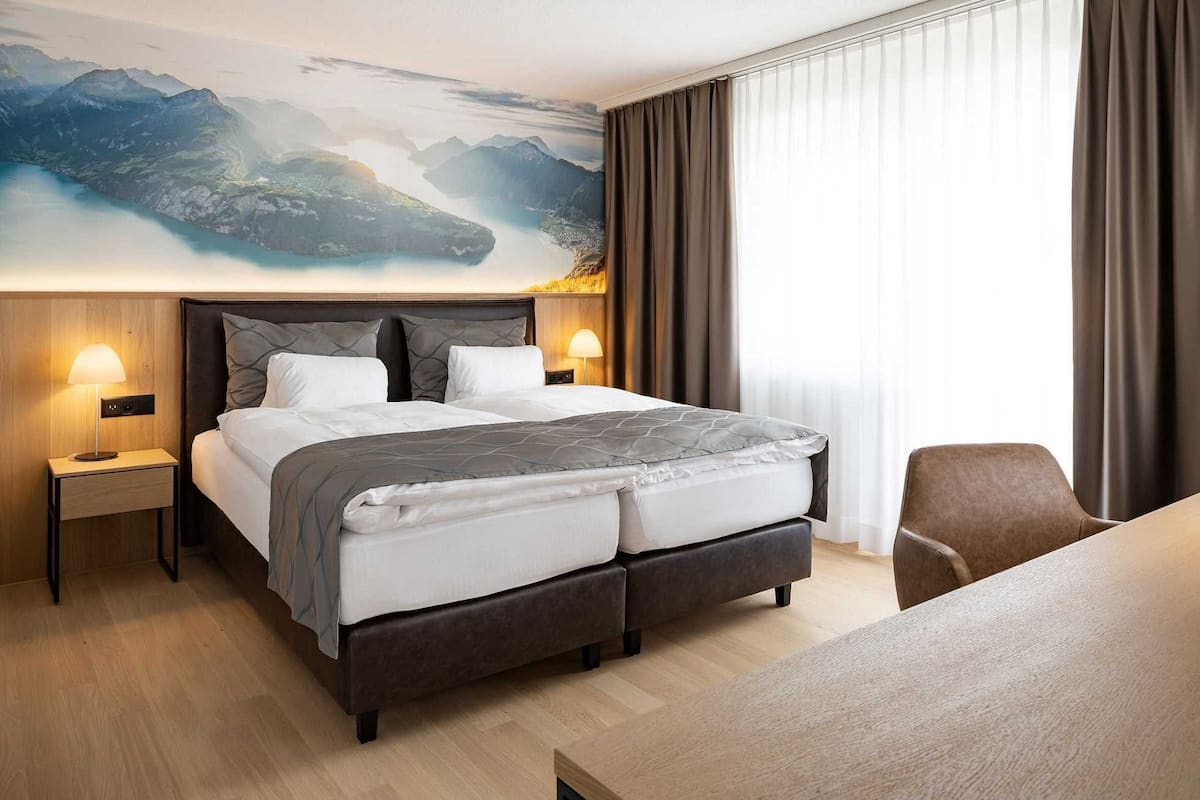 Habitación de hotel con una cama grande, dos mesitas de noche con lámparas, vistas a la montaña por la ventana y una mesa de comedor.