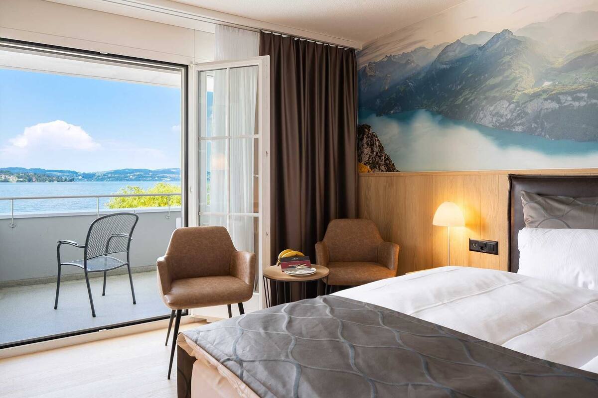 Habitación de hotel con cama, dos sillones, una mesita, una lámpara y un ventanal con vistas a un lago y montañas.