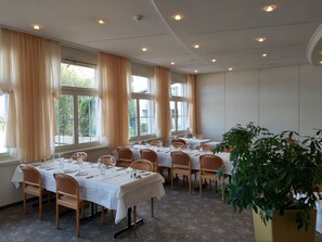A dining area with tables set for a meal, large windows with curtains, and a potted plant. - Seehotel Winkelried (Stansstad)