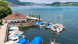A lakeside marina with boats, a restaurant with outdoor seating, and a backdrop of hills. - Seehotel Winkelried (Stansstad)