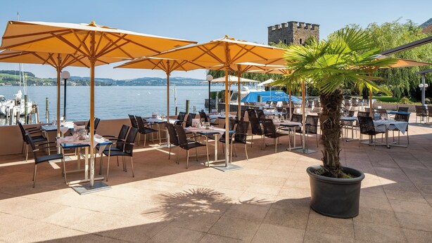 An outdoor dining area with tables and chairs, umbrellas, and a view of the water. - Seehotel Winkelried (Stansstad)