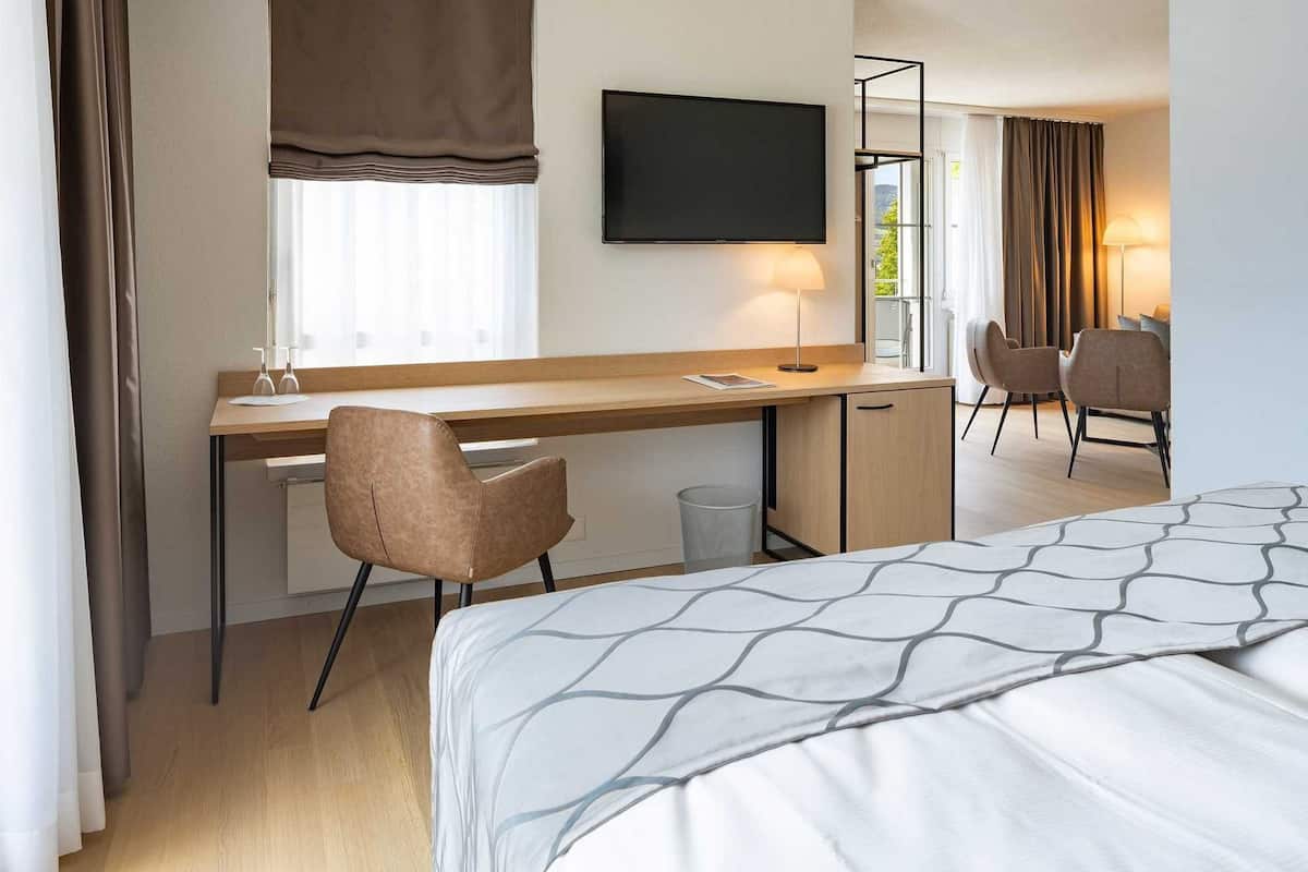 Una habitación de hotel con cama, un escritorio con silla, televisor y ventana con cortinas.