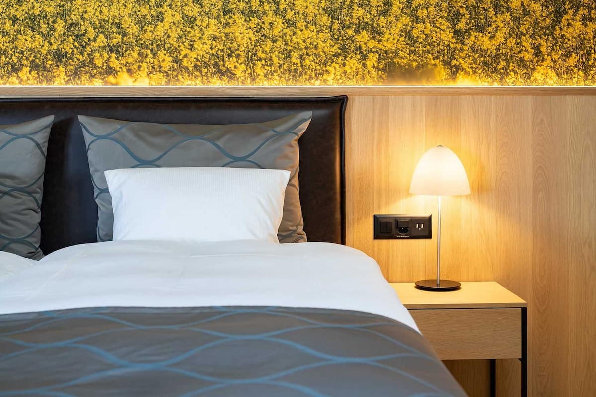 Una cama bien tendida con una colcha estampada, un cabecero con almohadones decorativos, una lámpara de noche y una pared con papel tapiz floral.