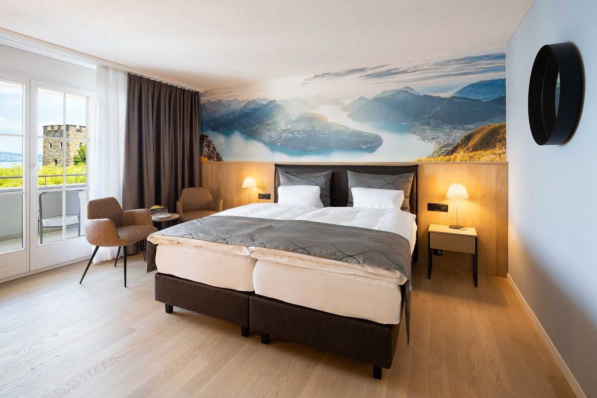 Habitación de hotel con una cama grande, dos mesitas de noche con lámparas, una silla y vistas a un paisaje montañoso a través de un ventanal amplio.