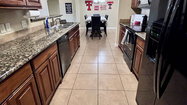 A kitchen with wooden cabinets, granite countertops, a black refrigerator, and a dining area with a table and chairs. - Raiderland Rest Stop - 3BR/2BA - Close to Campus (Lubbock)