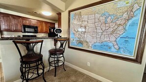 A kitchen with wooden cabinets, a microwave, and two bar stools. A large framed map of the United States is on the wall. - Raiderland Rest Stop - 3BR/2BA - Close to Campus (Lubbock)