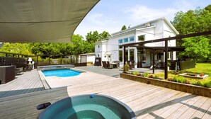 A wooden deck with a hot tub and a swimming pool, a covered patio area, and a white house in the background.